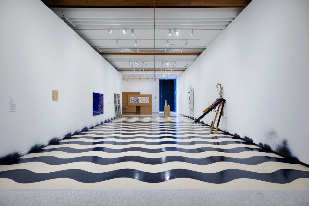 Andrew Barber in A world undone The image shows a long white corridor, looking from one end down to the other. On the floor of the corridor is a series of spray-painted black waves, which run horizontally down the length of the corridor. Black paint residue is smudged on the walls at the end of each of the waves. On the left hand wall is a small, light pink artwork hung high up from the floor, and further down is a large reflective dark blue square artwork. Another two tall sculptures are just visible leaning against the wall at the other end of the corridor. On the right wall, a white canoe hung vertically on the wall is just visible at the end of the corridor, and closer to the viewer is an easel opened out and leaning precariously against the gallery wall. At the end of the corridor a tall cardboard structure is visible but far away.