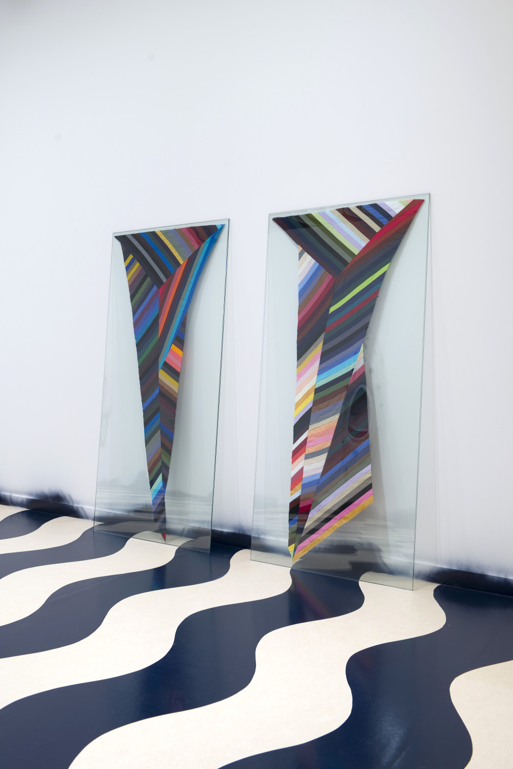 Amy Howden-Chapman in A world undone Two tall, vertical glass panes lean side by side against a white gallery wall. The floor below them is covered in a black and white wavy pattern. The glass panes have two similar but slightly different tapered designs sandwiched in between them, which are made up of thin strips of colour ranging from light pastel to dark blacks, including green, red, blue, purples, oranges, grey, and white in multiple shades. The strips are disjointed and formed trapezoid patterns within the design. The overall tapered effect in each of them is V-shaped.