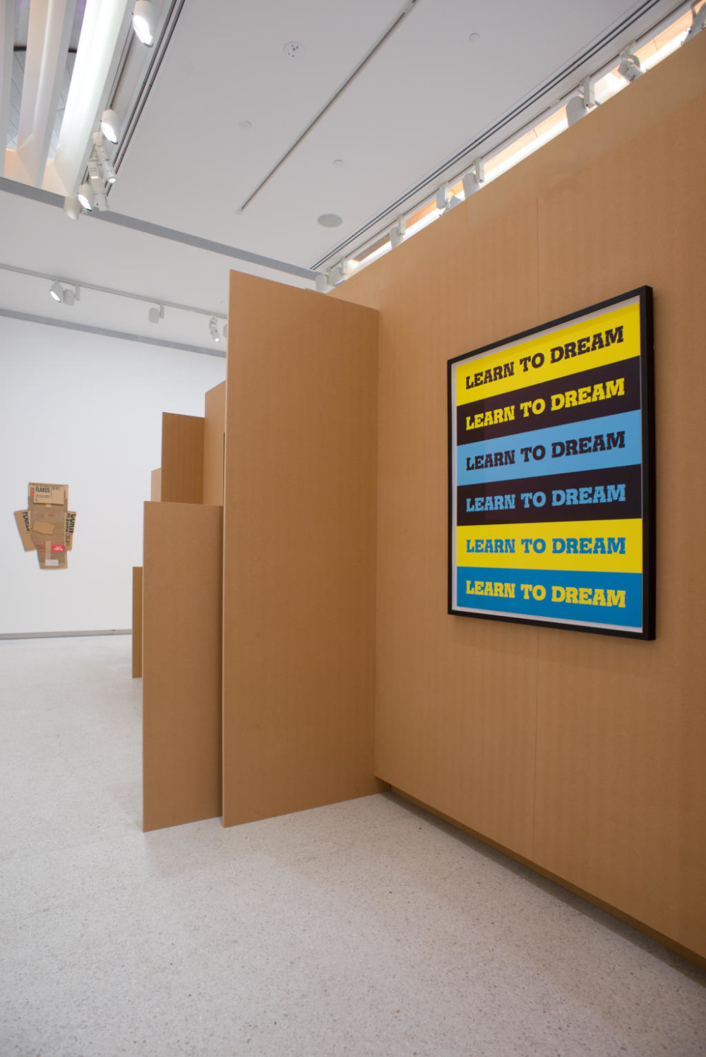 The image shows the other side of the freestanding cardboard structure. On a wall of the structure hangs a screenprint consisting of blue, yellow and black horizontal stripes, each of which has 'LEARN TO DREAM' written in capitals in black, yellow and blue on each stripe so it repeats across the entire print. On the wall of the main gallery space in the background, a 2D work made up of found carboard scraps haphazardly arranged together can be seen.