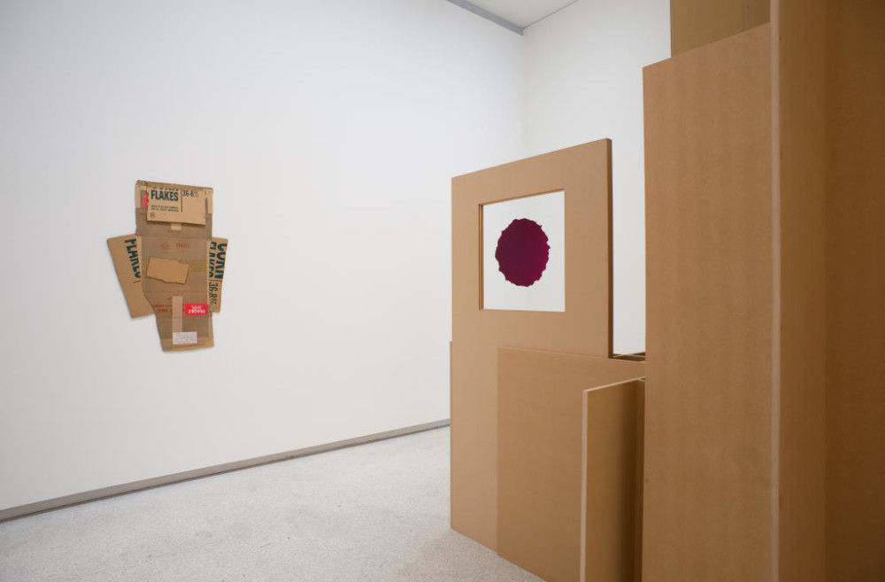 The image shows one edge of the freestanding structure. A square of white with a rough-edged purple circle in the middle of it is displayed on the edge of the structure. On the main gallery wall next to the structure hangs the 2D found cardboard artwork described in the last image; now it is clear that the work is made up of reused cereal boxes, as the lettering 'cornflakes' in a stylised font is clearly visible.