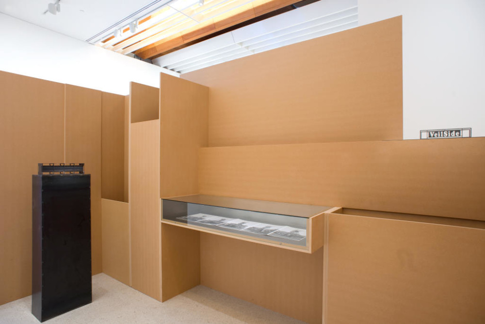 Another side of the freestanding cardboard structure is depicted. Here a tall, black sculpture shaped like a tall oblong is placed over to the left side. In the centre of the structure a horizontal glass vitrine has been built into the structure, with black and white photographs laying inside.