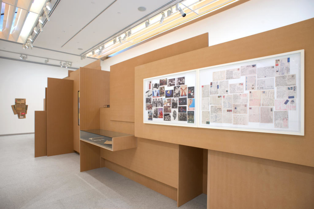 The image shows another part of the cardboard structure. On this side, another horizontal glass vitrine is visible in the left hand side of the structure, with two objects inside which aren't clearly visible. On the wall to the right of this vitrine hang two large framed collages of photographs and newspaper and magazine clippings arranged in a jumbled order.