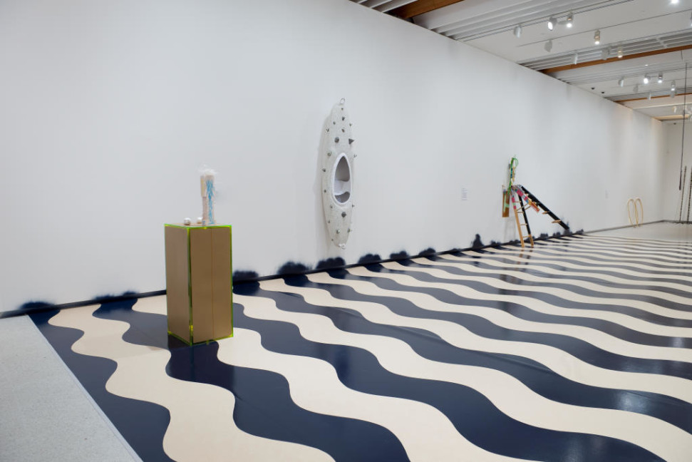 The image shows the corridor space from the other end of the room. The black and white waves ripple across the floor, and a tall narrow plinth edged in bright green with brown sides sits at the nearest end of the corridor with a smaller white painted figure perched on top of it. On the wall a little further down the corridor hangs a long white canoe flat on the wall, covered in large pieces of spiky dried paint which has been layered to produce a heavily 3D effect. These large masses are dotted all over the canoe hull. Further down the corridor, a small wooden stepladder leans against the wall, violently tilted so that its back legs are off the floor.