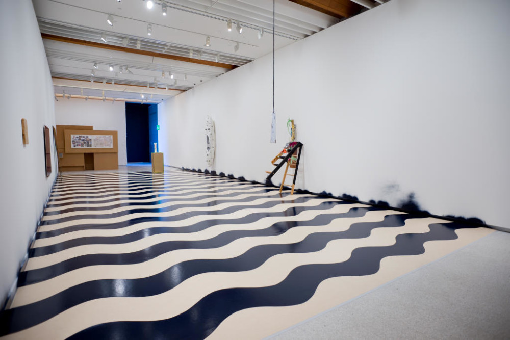 The image shows the other angle of the gallery corridor. The ladder leaning against the wall is visible again, as well as the wavy-patterned floor. In the distance the canoe is visible and so is the cardboard structure at the end of the corridor space. In the background of this is a doorway into another room, painted in a dark black colour with blue light emitting from it. 