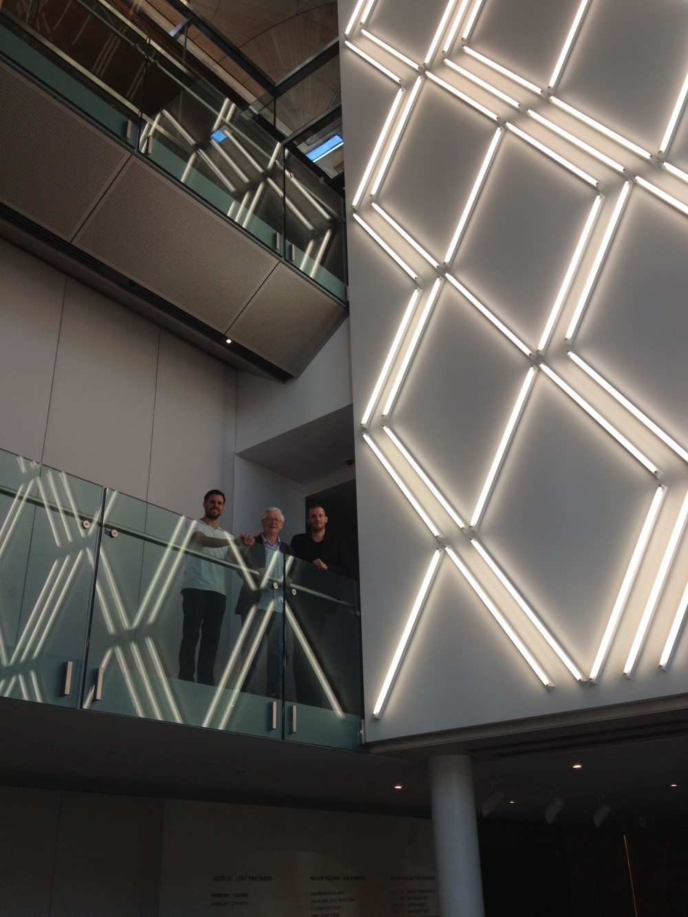 Three men stand on a glass lined balcony in the atrium of Auckland Art Gallery, right next to the bottom section of Jonathan Jones' huge glowing work as described in the previous image. The glowing tubes are reflected on the glass surface of the balcony. 