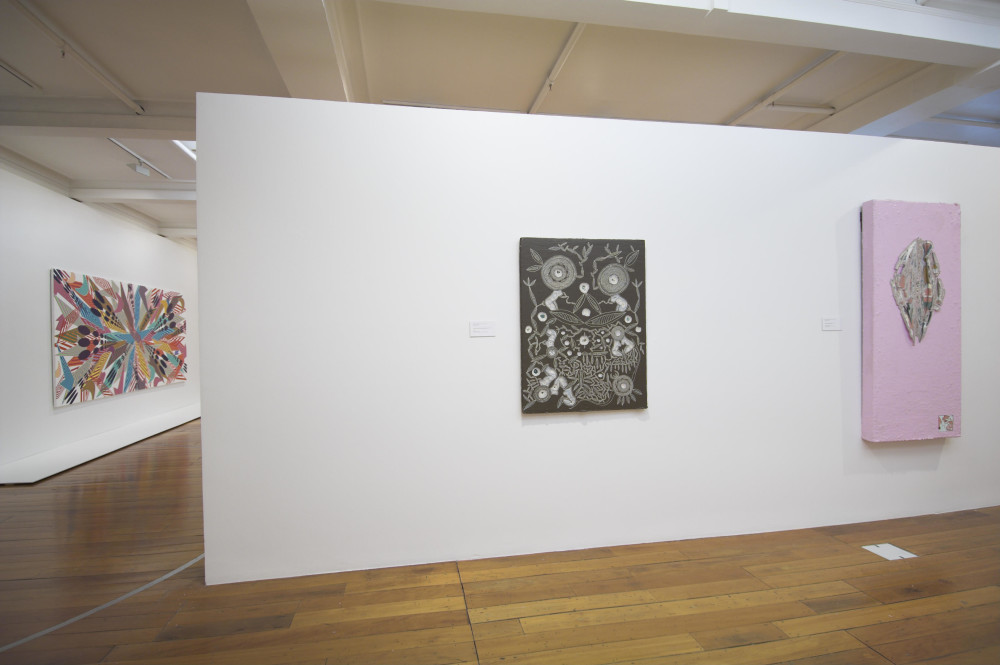 The image shows a white gallery space with a polished wooden floor. In the background on an opposing wall hangs the large horizontal painting described in the image preceding this one, with all multicoloured patterns converging into a single point. On the wall at the front of this image, on the left hand side, hangs a brown portrait painting designed to look like carved wood. Multiple circular patterns have been carved out of the surface of the painting, showing circular patterns with small, disembodied female figures arranged in a variety of poses in different patterns. On the right hand side of the wall is a light pink, portrait painting with a slit in the middle of it, and multiple layers of different coloured paint peeled away to expose a crevice in the work.