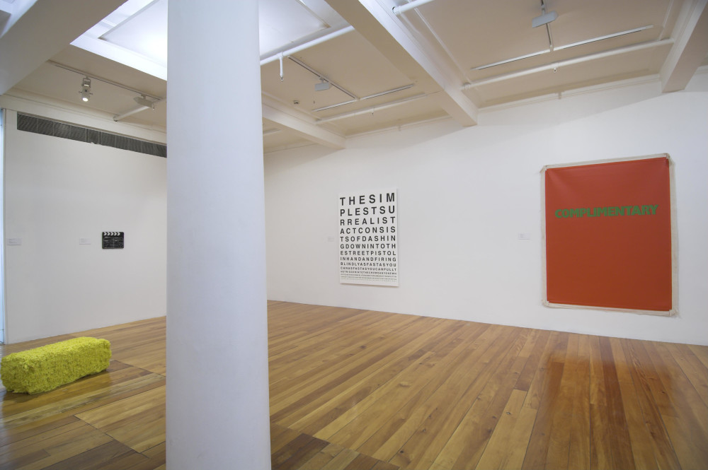 The image shows a white gallery space with a polished wooden floor. In the immediate foreground of the image is a large white column which cuts through the image. On the left hand side of this column is an unnaturally yellow work on the ground of the gallery, which resembles a bale of hay. On the wall above it is a director's clapboard, and on the right hand side of the column is a large vertical artwork with a series of phrases laid out on it in the manner of an optometrist's eyechart. The letters start off huge and reduce in size as the lines progress down the canvas. Next to this artwork, on the far right is a large almost square bright red work with green text on it. 