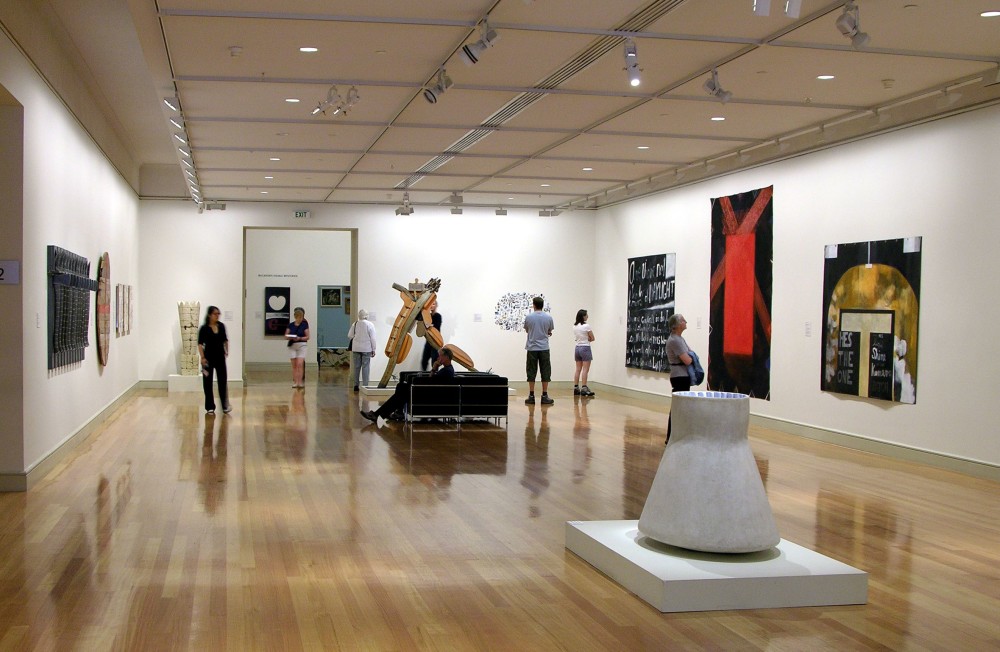 The image shows a white gallery space with a polished wooden floor. In the centre of the room, a sculpture of a grey concrete triangular shape sits on a flat plinth. On the walls around the sculpture hang multiple artworks of various sizes and colours, with people standing around in the space contemplating them. 