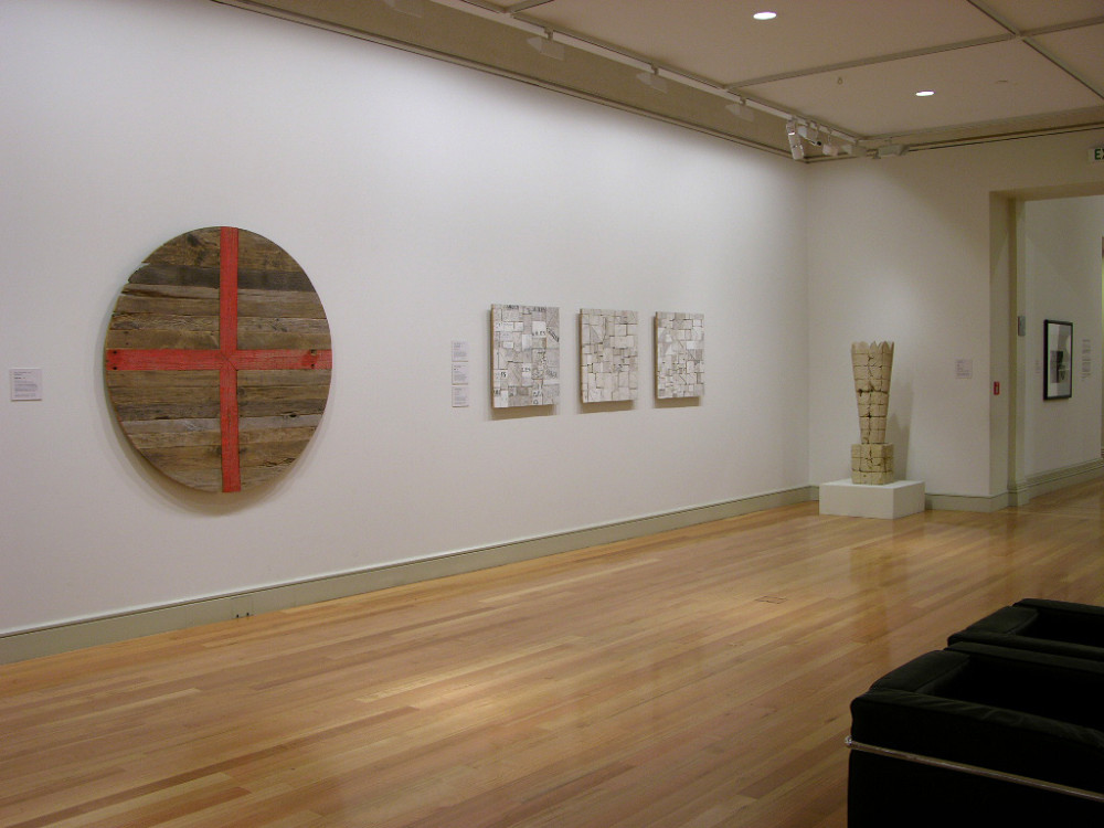 The image shows artworks hanging on a wall. On the left hangs a large circle of wood with a bright orange cross painted through it. Next to this hang three small artworks comprised of lots of small pieces of chopped wood arranged together and whitewashed. On the right stands a large abstract sculpture which tapers outwards. 