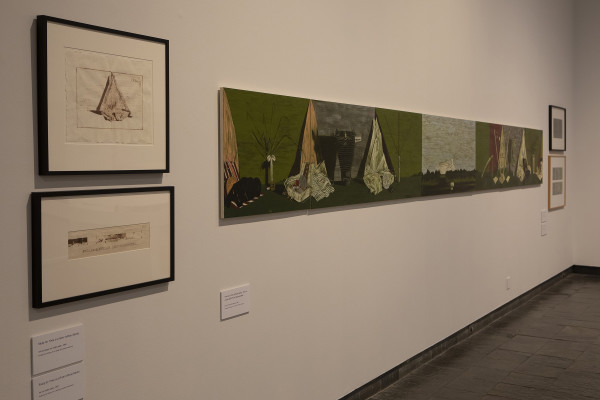 The image shows a gallery wall in Waikato Museum. On the left are two drawings in square frames which appear to be studies for the main, large painting next to them. The large painting is a long horizontal work stretching away from the viewer, showing objects arranged in still life compositions against light green backgrounds with landscapes in between.