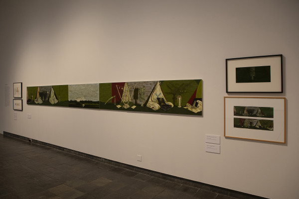 The image shows a gallery wall in Waikato Museum. On the left is a long horizontal painting stretching away from the viewer, showing objects arranged in still life compositions against light green backgrounds with landscapes in between. On the right are two framed studies for the work. 