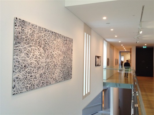 Emily Kame Kngwrreye A view down the corridor of one of the gallery spaces at Auckland Art Gallery, second floor. On the left wall hangs an artwork with a black background filled with white squiggly lines. Gallery visitors can be seen walking around in the distance.