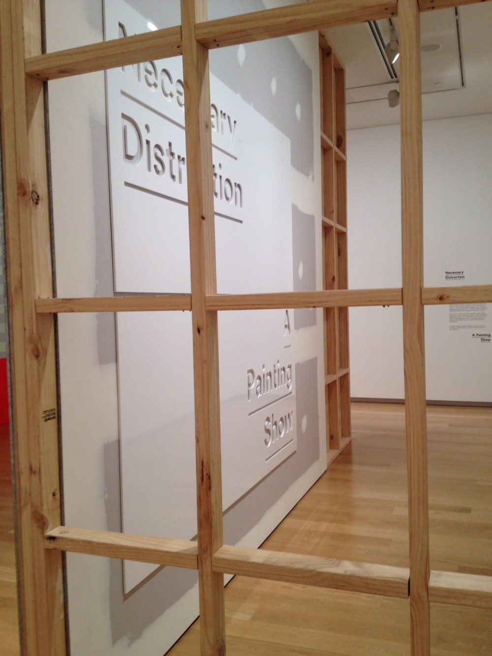 An image of the first wall of the exhibition. A wooden grid is in the foreground, and through it an unpainted gib board wall can be seen with a large white sign mounted on it. The sign reads 'Necessary Distraction, A Painting Show'. 