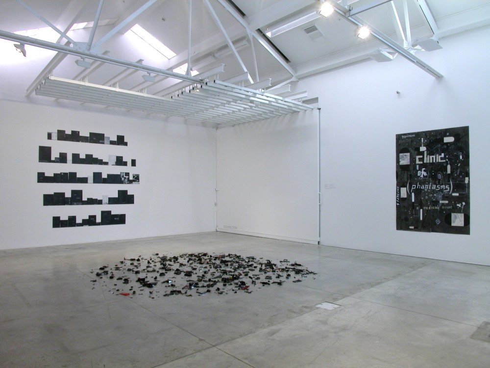 The image shows a white gallery space with a concrete floor. On the left hand wall is an artwork consisting of multiple horizontal rows of small books, which are all varying heights and widths. The shelves are arranged in so that the edges of the entire installation look rounded off in an almost circular shape. The books are a mixture of black, grey and white and have indiscernible words on them. On the floor in the centre of the room is a work which consists of lots of little, miscellaneous mechanical parts in black and silver scattered haphazardly around in a circular arrangement. On the righthand wall is a large black, vertical artwork which has rectangular shapes in dark grey scattered across it, with the words 'clinic of phantasms' written in the centre. 