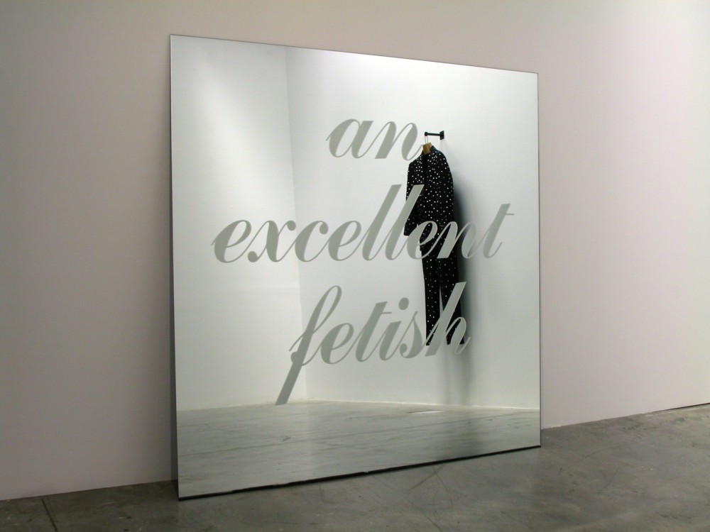 The image shows a square mirror leaning against a wall with the words 'an excellent fetish' engraved upon in it cursive writing. A black men's suit covered in little metal studs, hanging from a wall is visible in the reflection of the mirror. 