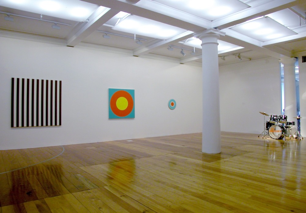 The work depicts the corner of a white gallery room with a polished wooden floor. On the left hand wall hang three artworks. The left work is a large square painting divided into even vertical strips of black and white. Next to it is a square artwork which consists of a small yellow circle within a larger orange circle, on a light blue background. To the right of this artwork hangs a much smaller circular artwork, which shows a small orange circle inside a bigger white circle inside a bigger light blue circle. Against the right hand gallery wall a full drum kit has been set up, with the words 'The Anguses' written in capital orange letters across the front of the largest drum. 