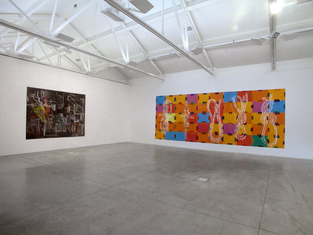 The image shows a corner of a white gallery space with a concrete floor. On the left hand wall hangs a large horizontal painting covered in messy, angular abstract white and grey lines, with multicoloured scribblings, against a red and black cloudy background. On the right hand wall hangs a very long oblong, large work consisting of tiles of blue, orange, red, purple and green with different kinds of knot diagrams painted over the top in white. 