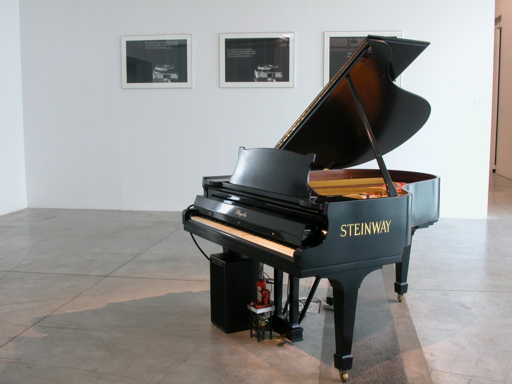 The image shows a white gallery space with a concrete floor. On the back wall hang three framed artworks, each with a circular slide holder in the foreground and small, indiscernible white text printed next to them. In the centre of the room is a large Steinway grand piano. 