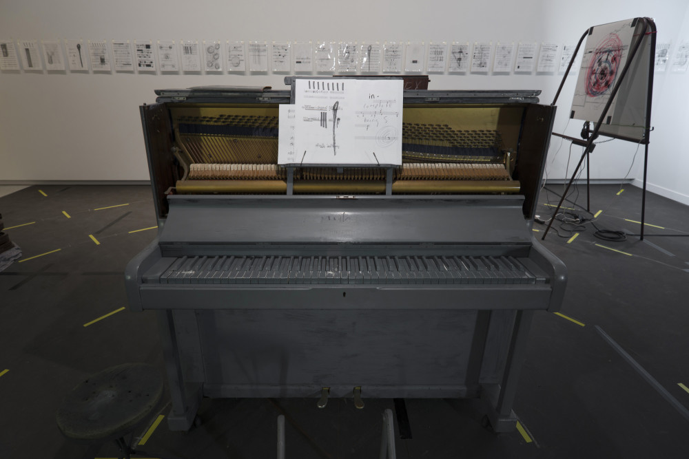 Shout Whisper Wail The image shows the same installation depicted in the previous image, but taken from the other side. In the centre of the image stands a large upright piano painted entirely in grey paint, with sheet music propped up on a stand above the keys. The sheet music is fragmented and has scrawls and scribbles all over it. Around the piano on the floor are the yellow off-cuts of tape circling the room, while on the wall behind the piano are multiple small drawings and prints in plastic protectors all in a row. To the right of the piano is one of the easels holding a large print.
