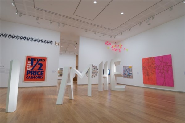 One of the ground floor galleries at Auckland Art Gallery is depicted. In the centre of the room large white three-dimensional lettering spells out 'I AM HE'. To the left hangs a red and blue work which reads '1/2 price cash only' on loose canvas. Above it, multiple identical signs bearing the letter 'O' are hung together on an angle. On the right wall hangs a pink and orange work with a smaller blue and yellow work next to it, and in the top right hand corner is a cluster of pink squiggly acrylic shapes.