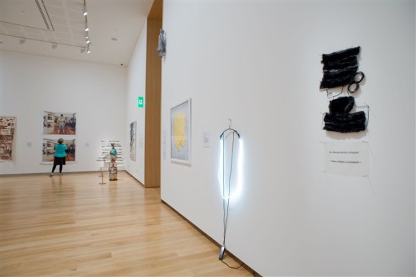 Multiple artworks hanging on the walls of a ground floor gallery space at Auckland Art Gallery. A woman stands in front of two loosely hung large photographs on the far wall of the gallery, looking at them. Next to her is a figurative sculpture of a person. On the wall closest to the viewer, a small grey figure of a business man hangs facing the wall, high above visitors. Below the figure is a framed painting. On the right of the framed painting, a coat hanger draped with bright white coils emitting light hangs from the wall. On the far right of this work, two ambiguous shapes wrapped in dark black wool are hung on the wall.