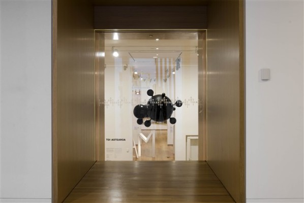 The image looks through a tall window on the mezzanine floor of Auckland Art Gallery. The window looks into the ground floor gallery, where a work consisting of multiple large black spheres connected to smaller black spheres hangs from the ceiling. 